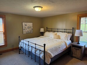 A bedroom with a metal bed frame, a bedside table with a lamp, a ceiling light fixture, and a window with blinds. - 5BR Farmhouse Lakeview • Wine Trail • Cozy Porch (Dundee)