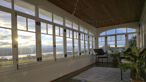 A spacious room with large windows offering a view of a lake and hills. There are two chairs and a rug on the floor.