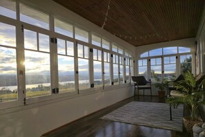 A spacious room with large windows offering a view of a lake and hills. There are two chairs and a rug on the floor. - 5BR Farmhouse Lakeview • Wine Trail • Cozy Porch (Dundee)