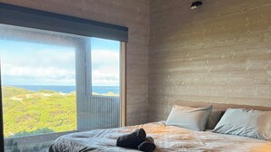 A bedroom with a large window offering a view of a green landscape and the sea. - Welcome to Admiral Cabin, a luxurious off-grid retreat on King Island, Tasmania. (Nugara)