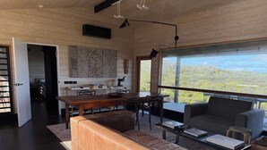 A spacious living area with a large window offering a scenic view, a cozy seating area, and a dining space with a wooden table and chairs. - Welcome to Admiral Cabin, a luxurious off-grid retreat on King Island, Tasmania. (Nugara)
