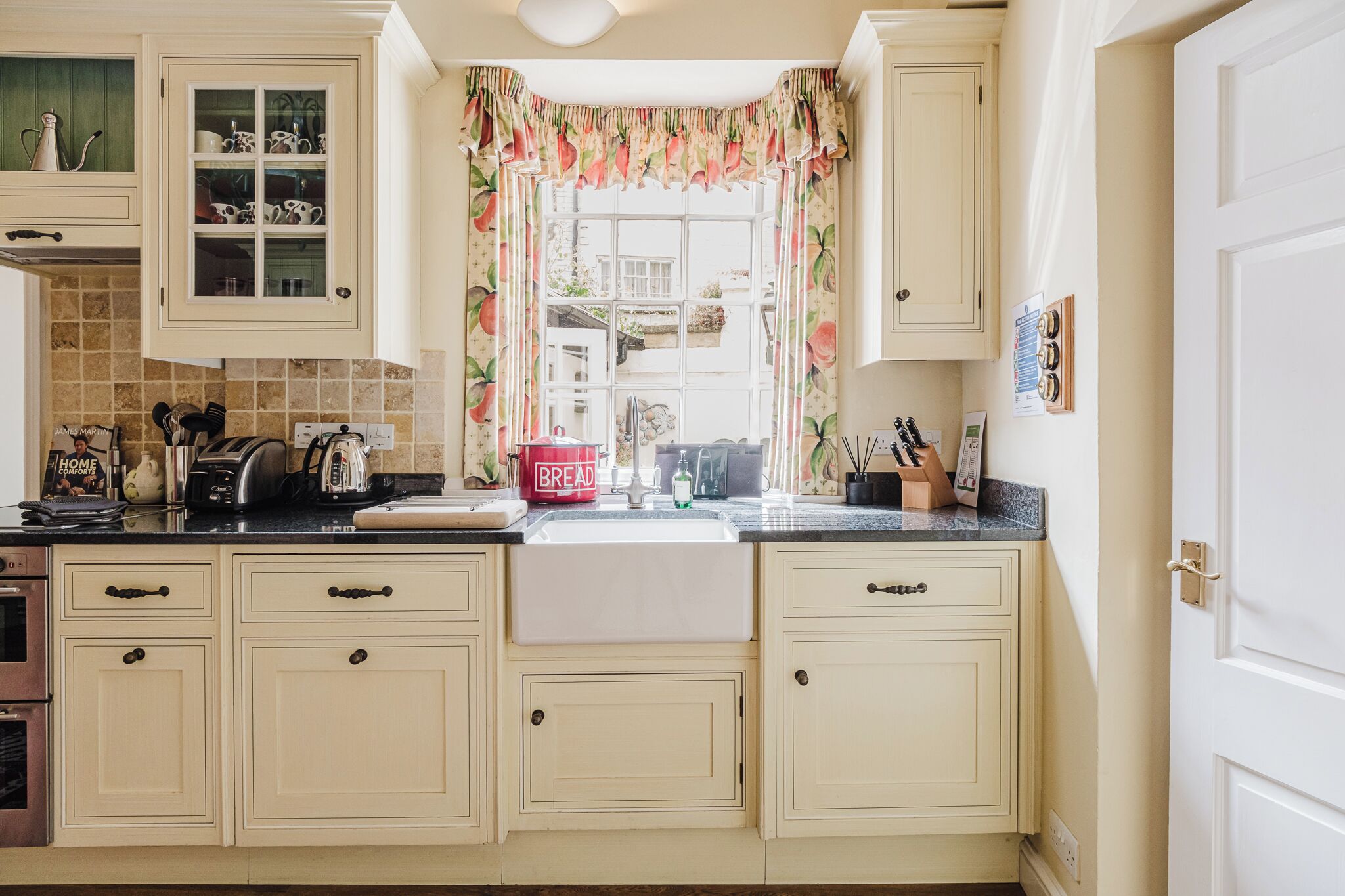 Une cuisine avec des meubles de couleur crème, un évier blanc et une fenêtre ornée de rideaux à motifs floraux.