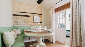 Un comedor con una mesa redonda dispuesta para cuatro personas, un banco verde, una pared de piedra y una puerta que da al exterior.