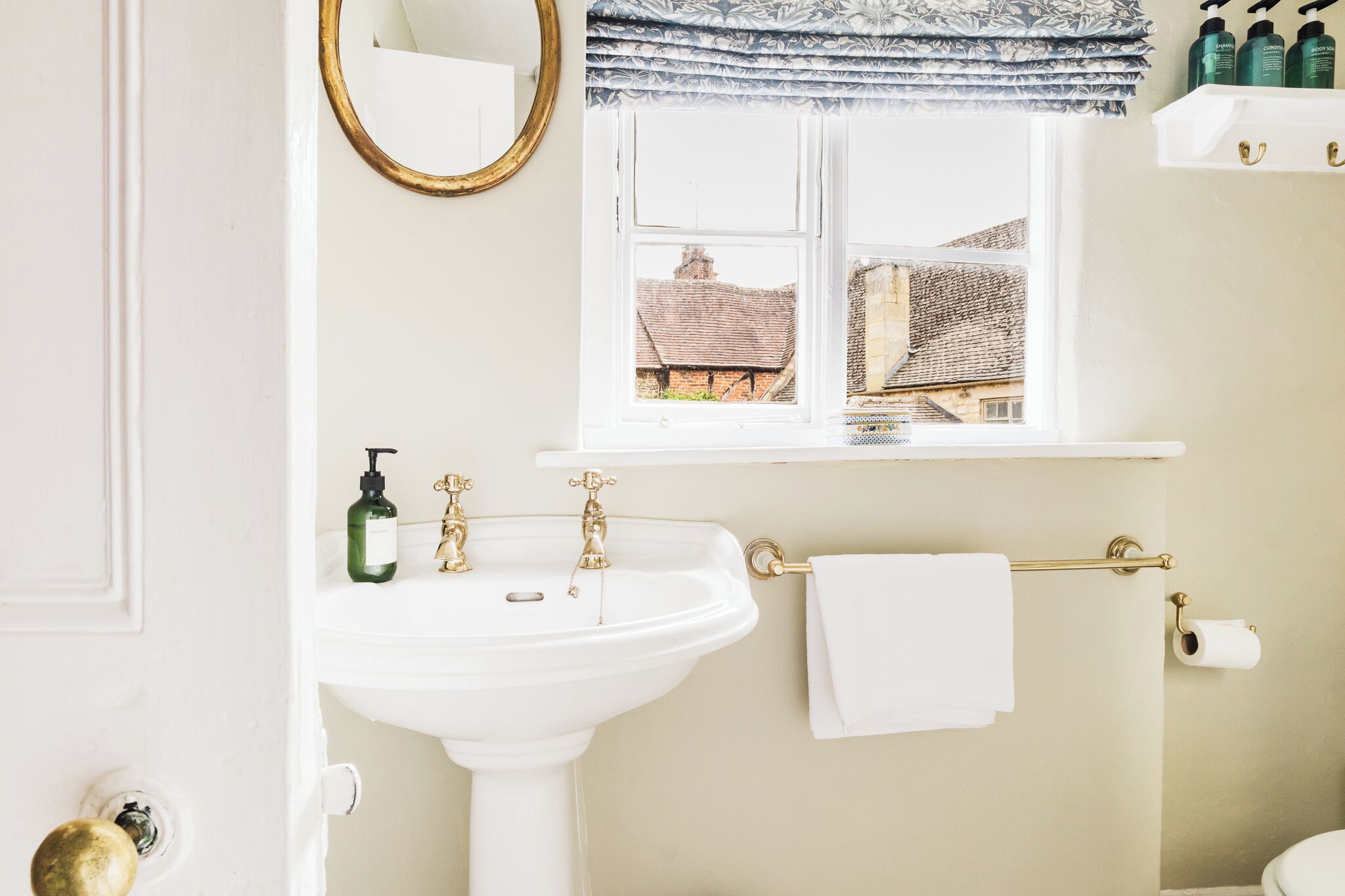 A bathroom with a white sink, gold faucets, a window with a view of buildings, a mirror, and toiletries on a shelf.