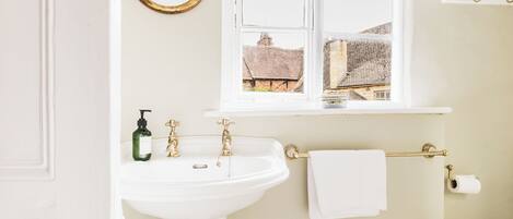 A bathroom with a white sink, gold faucets, a window with a view of buildings, a mirror, and toiletries on a shelf.