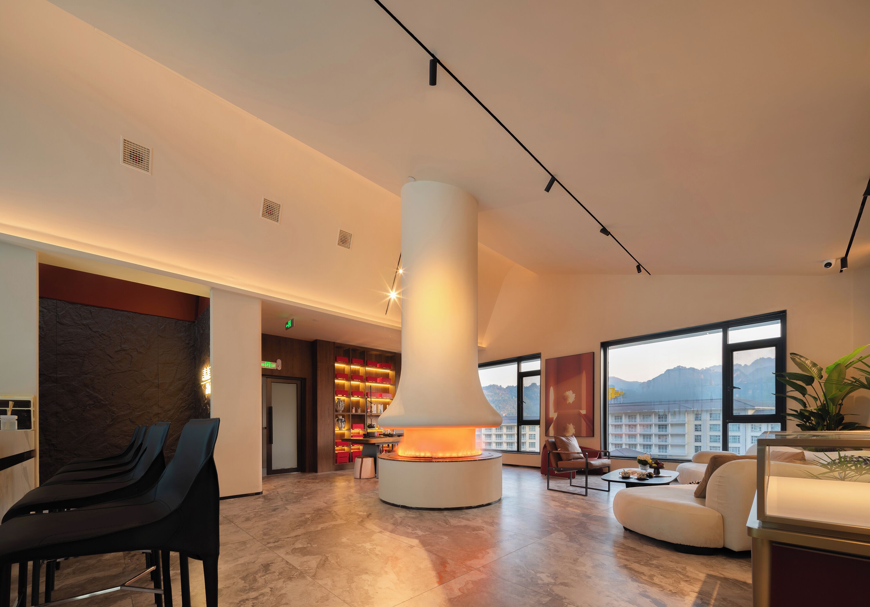 Un moderno vestíbulo de hotel con una chimenea central, zona de estar y grandes ventanales que ofrecen vistas a la montaña.