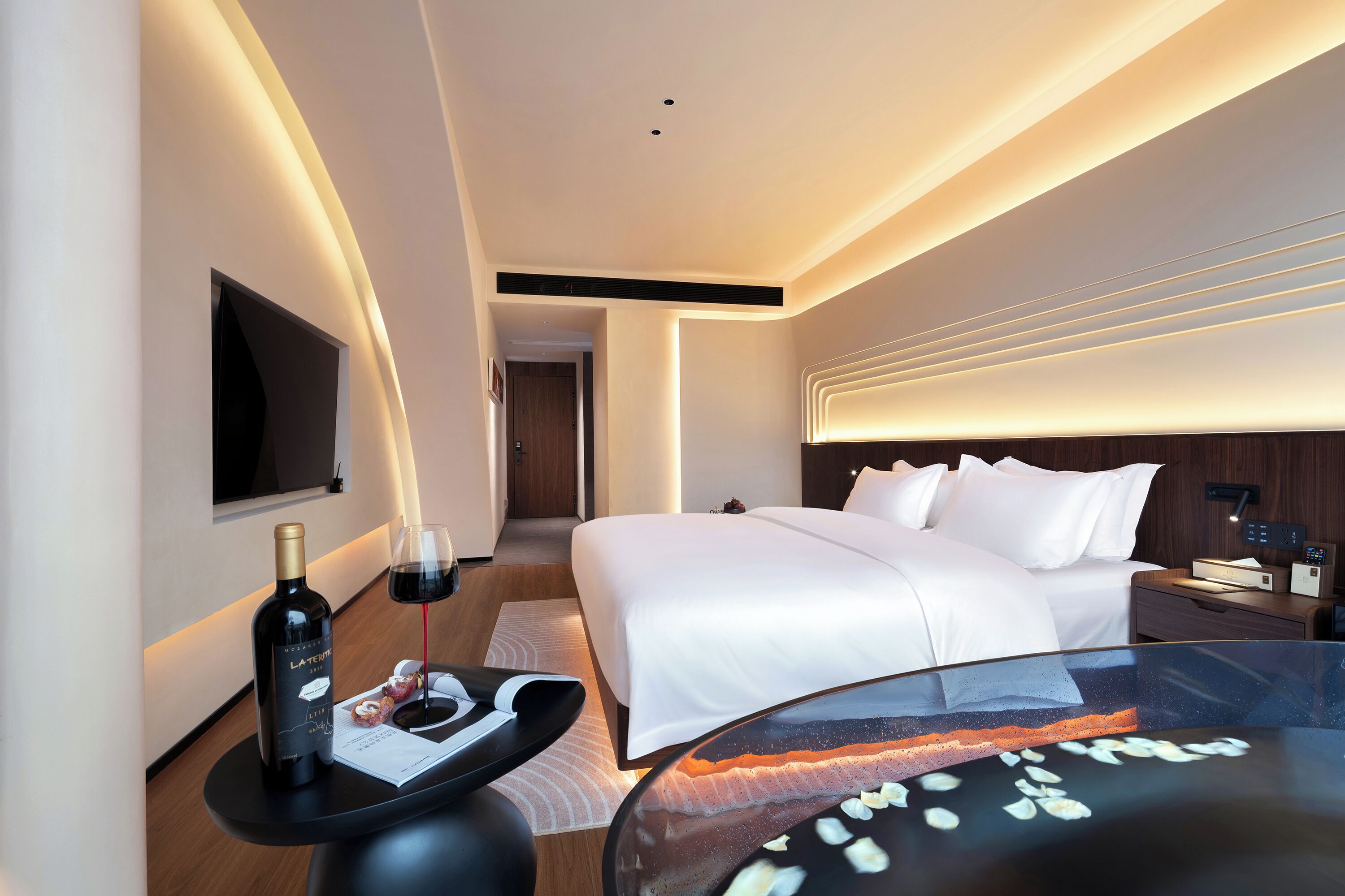 Une chambre d’hôtel moderne avec un lit soigneusement fait, un téléviseur à écran plat fixé au mur, une table de chevet avec une bouteille de vin et des verres, et une table en verre ornée d’un objet décoratif.