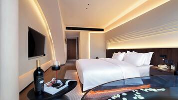 Une chambre d’hôtel moderne avec un lit soigneusement fait, un téléviseur à écran plat fixé au mur, une table de chevet avec une bouteille de vin et des verres, et une table en verre ornée d’un objet décoratif.