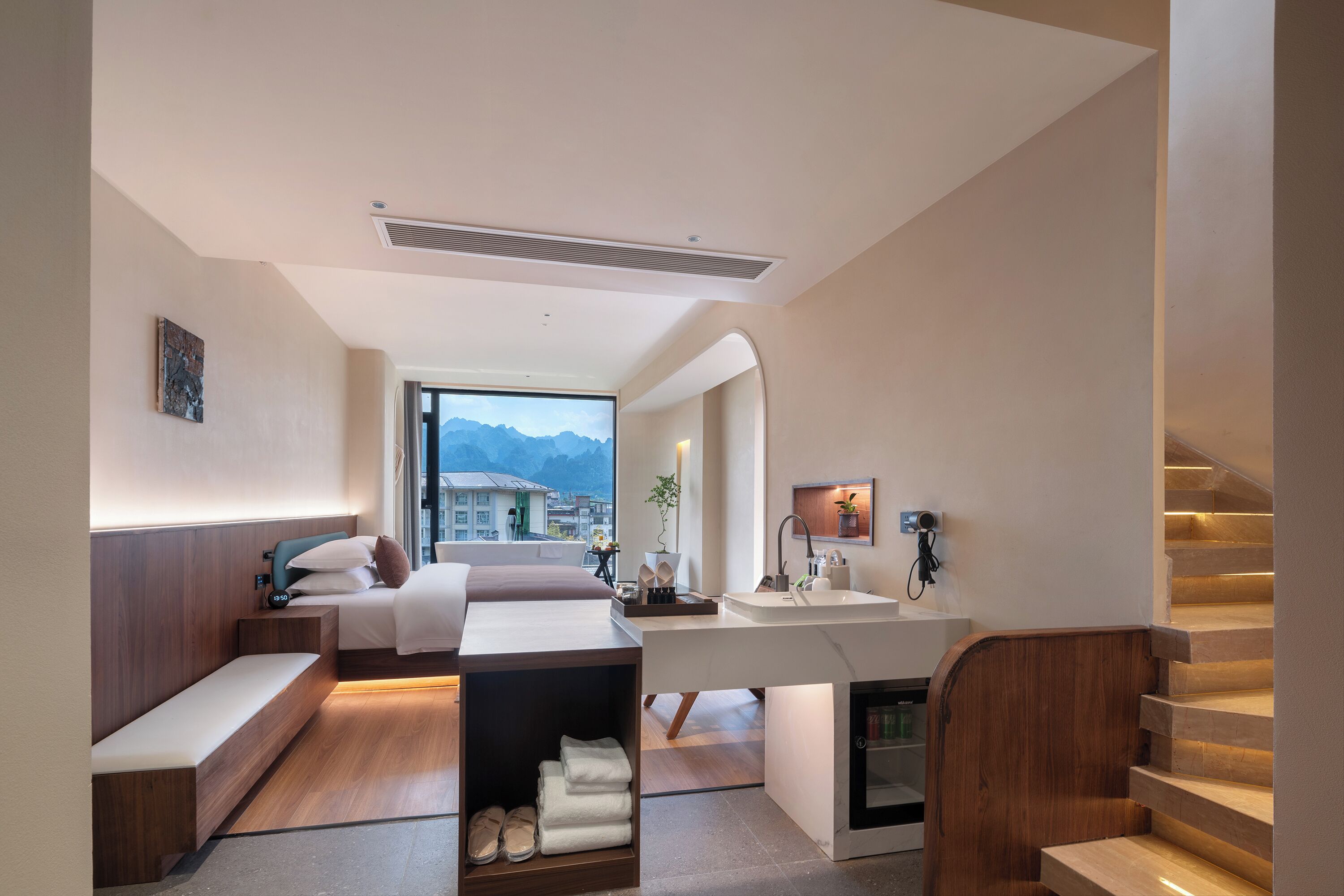 A modern hotel room with a bed, bedside table, bench, and a view of mountains.