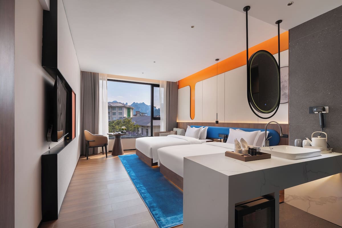 A hotel room with two beds, a large window, a desk with a sink, and a view of buildings and mountains.