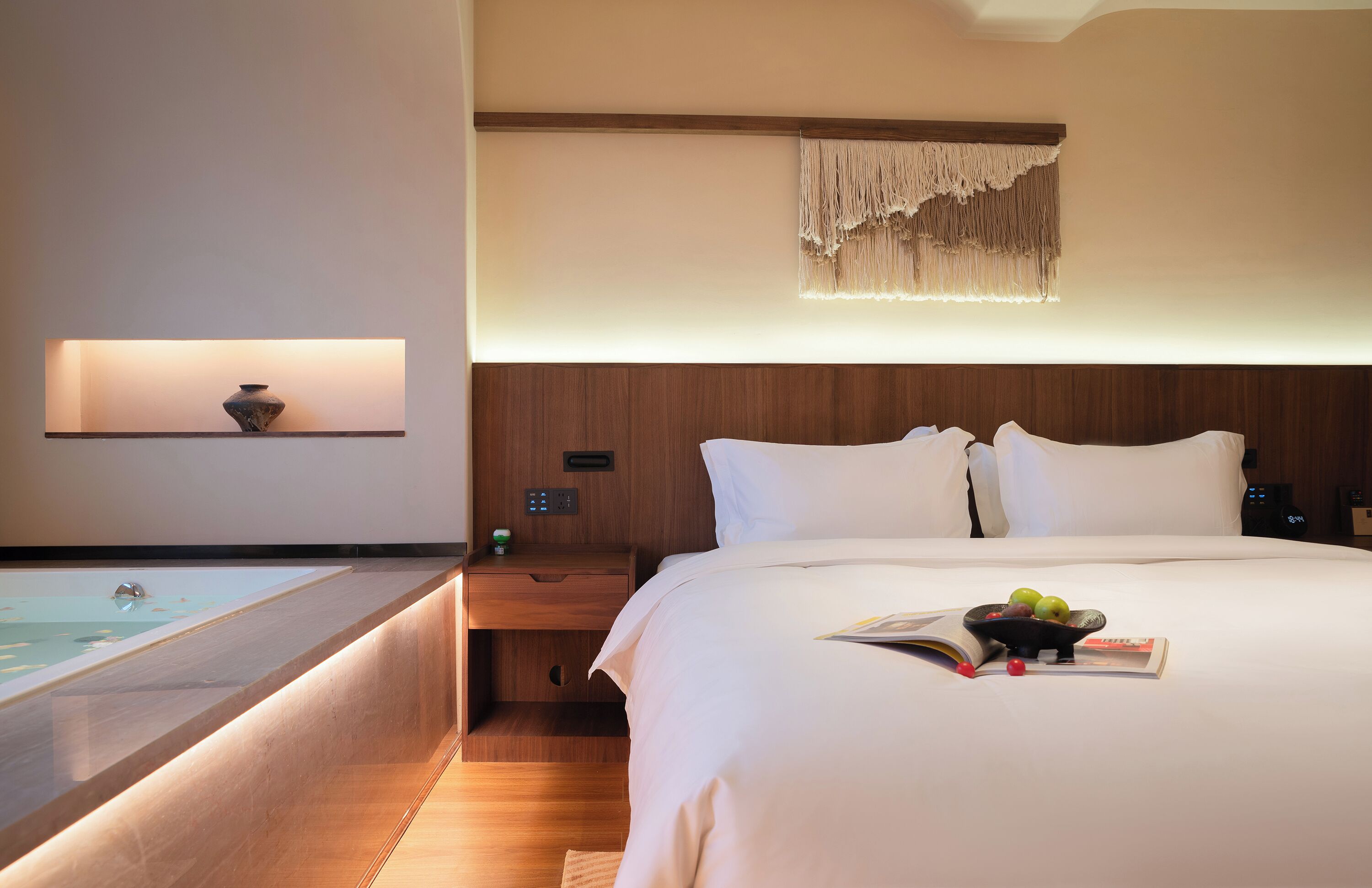 A hotel room with a large bed, a bedside table with a lamp, a small bedside table with a vase, a wall-mounted macrame piece, and a view of a pool.