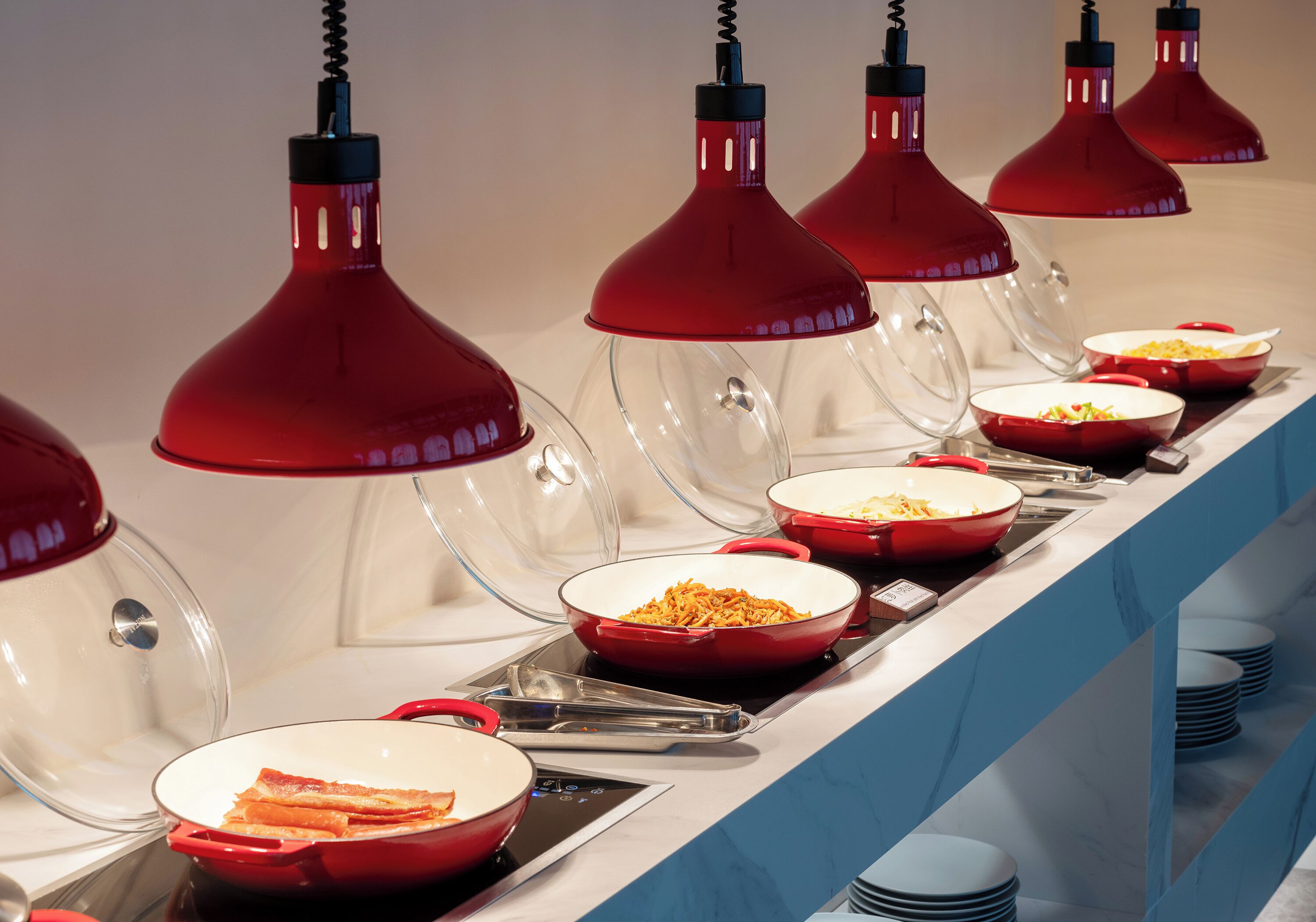 A buffet setup with multiple red bowls and clear glass covers, illuminated by red pendant lights.