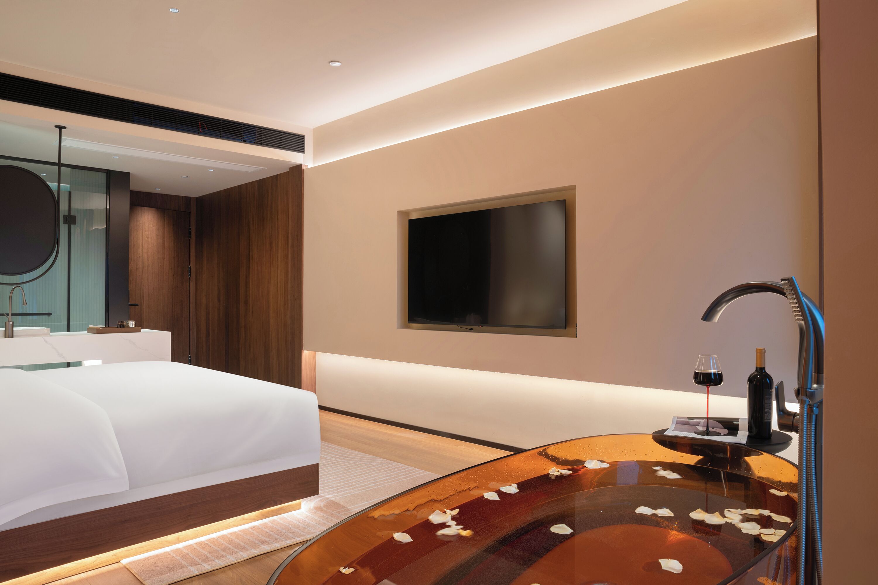 Quarto de hotel moderno com uma cama grande, uma banheira de vidro com pétalas de rosa, uma TV montada e uma garrafa de vinho.