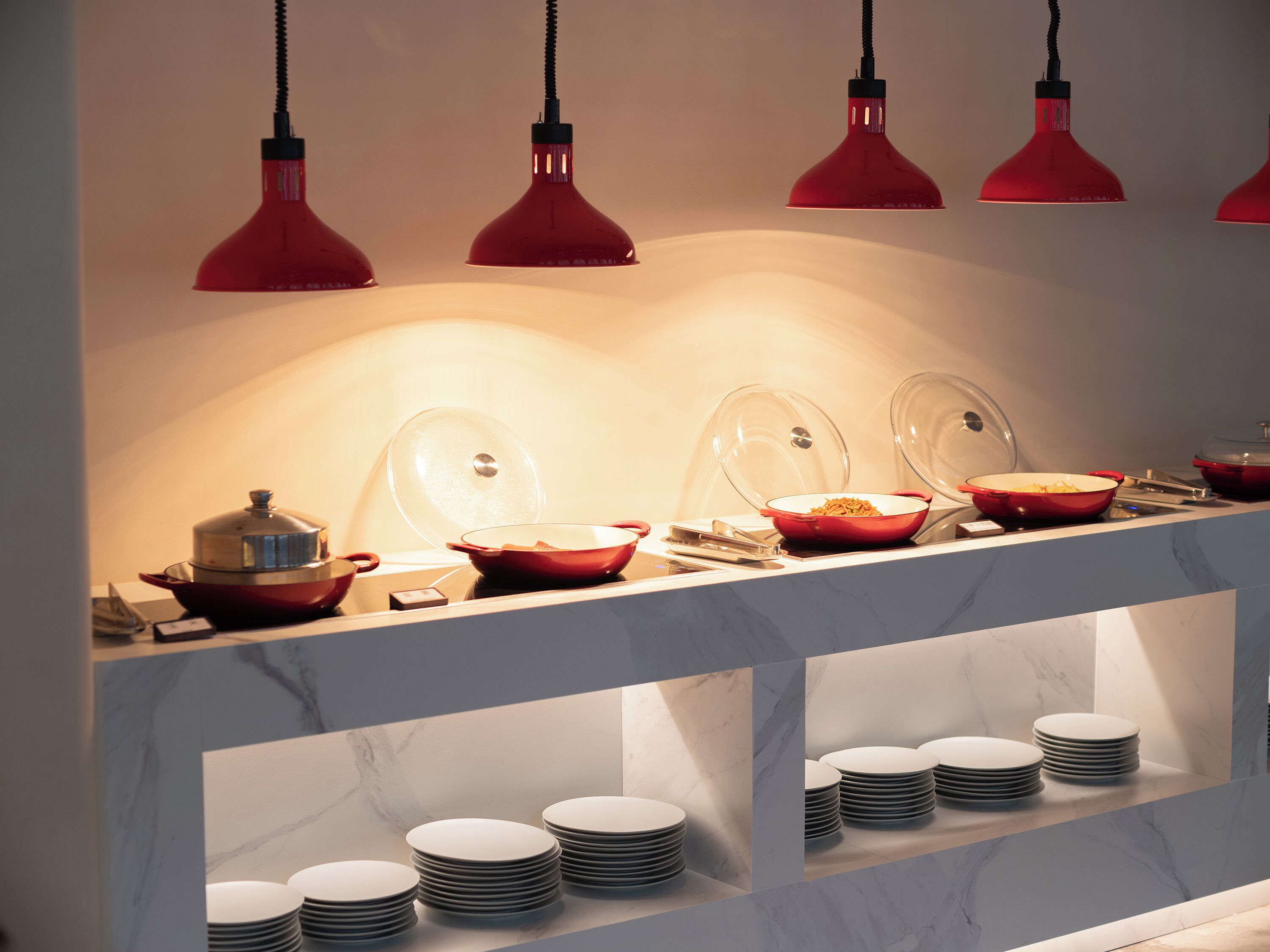 A buffet setup with multiple bowls and plates, overhead red pendant lighting, and marble countertops.