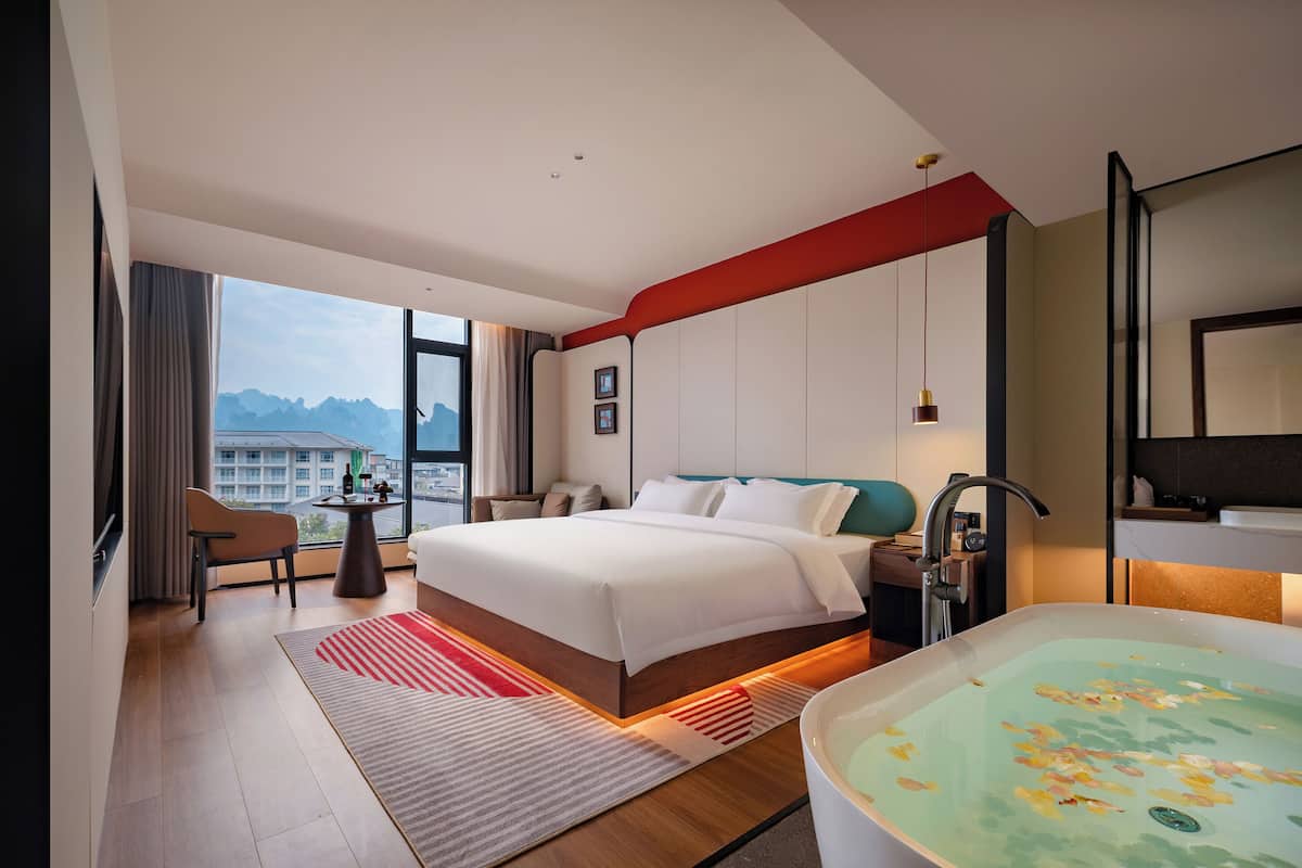 A modern hotel room with a large bed, a sitting area with a chair and table, a bathtub with floral design, and a city view.