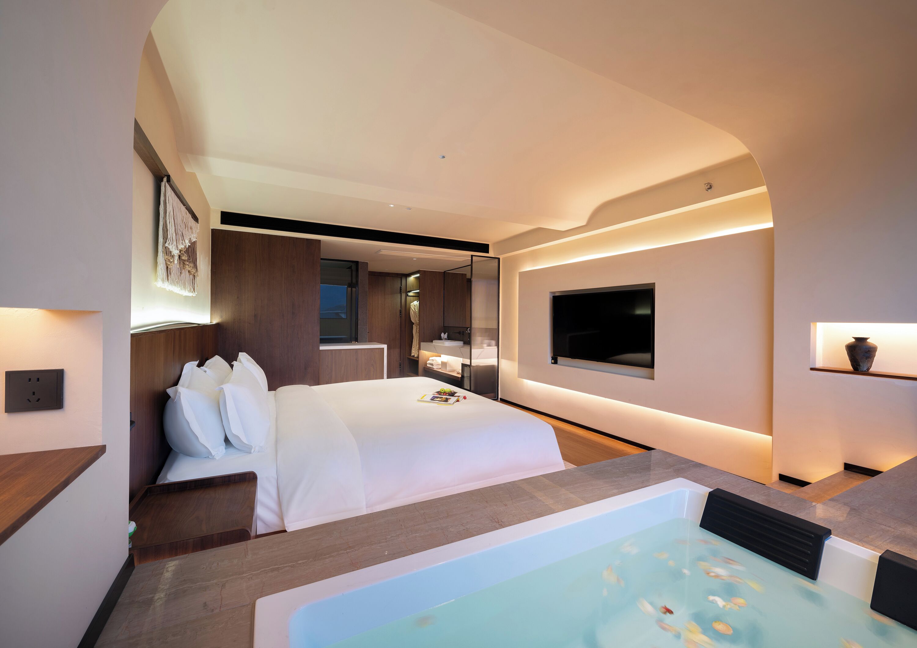 A modern hotel room with a large bed, a TV mounted on the wall, a bathtub, and a small window.
