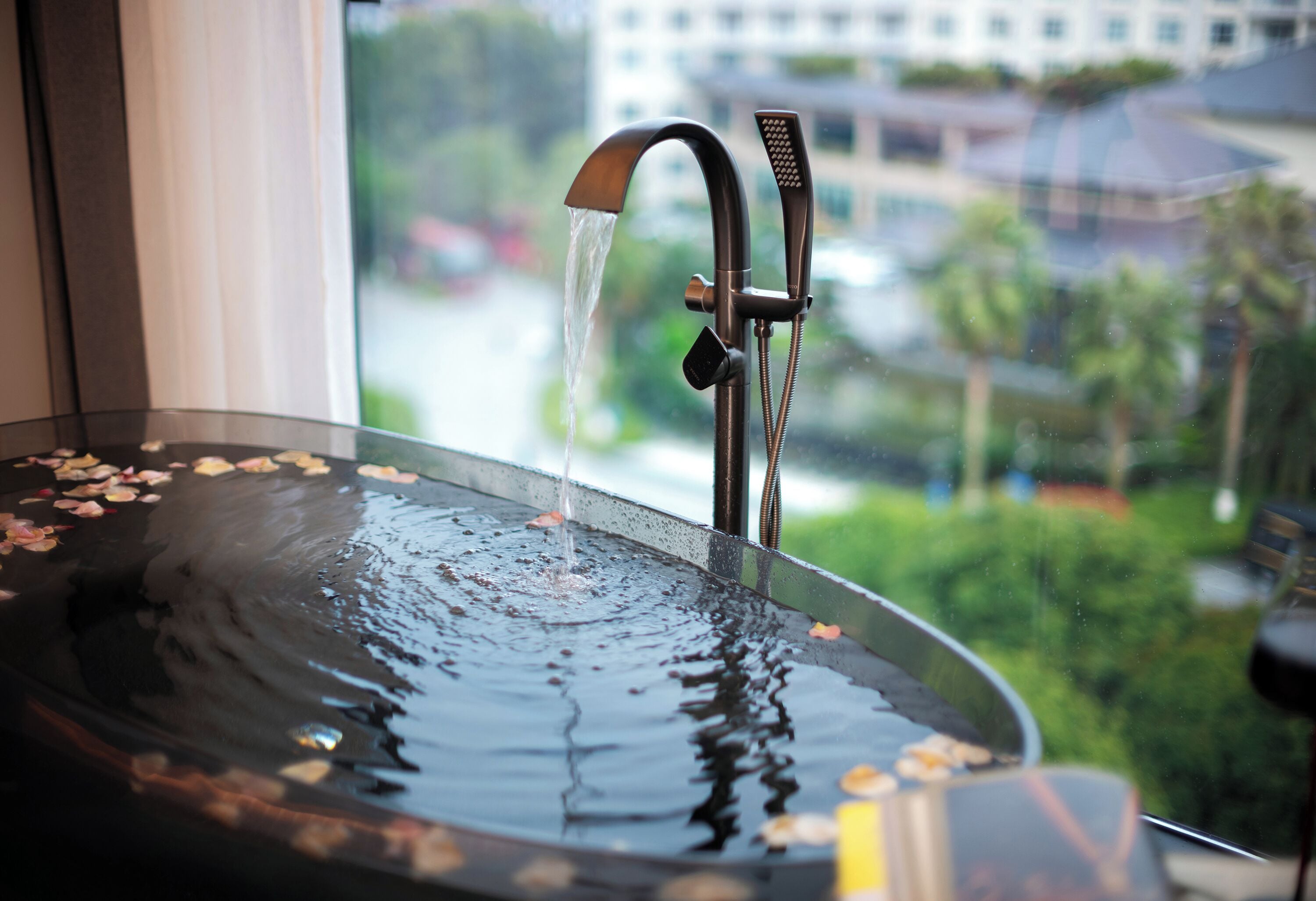 Une salle de bain dotée d’un élément décoratif aquatique entouré de verre, d’une pomme de douche et offrant une vue sur le paysage urbain par une fenêtre.