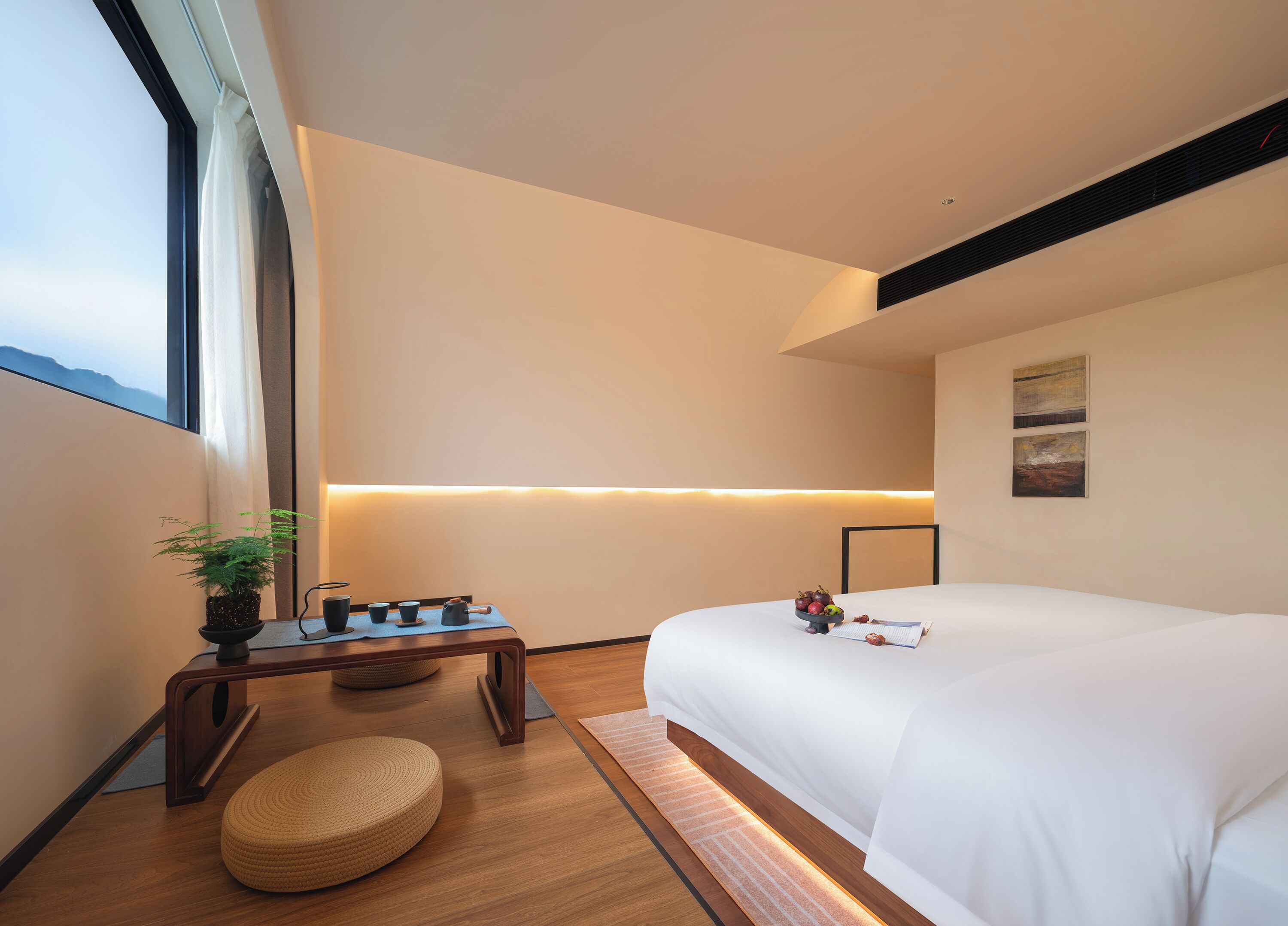 Ein modernes Hotelzimmer mit einem großen Bett, einem Nachttisch mit einer Pflanze, einem kleinen Tisch mit Tassen und einem Wasserkocher sowie einem Blick ins Freie durch ein großes Fenster.