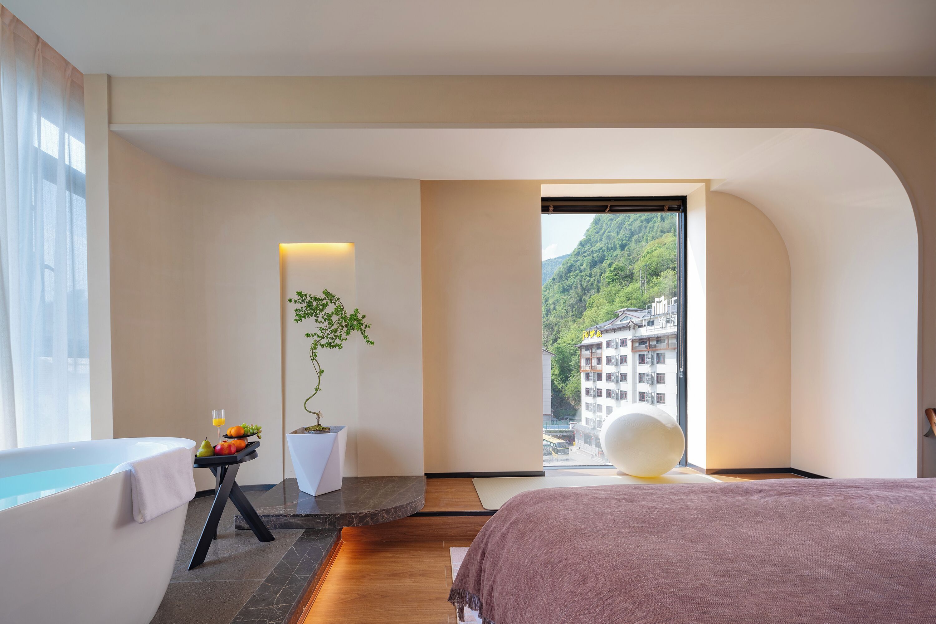 A modern bedroom with a bathtub, a bed, a small table with fruits and a glass, and a view of a mountain and buildings.