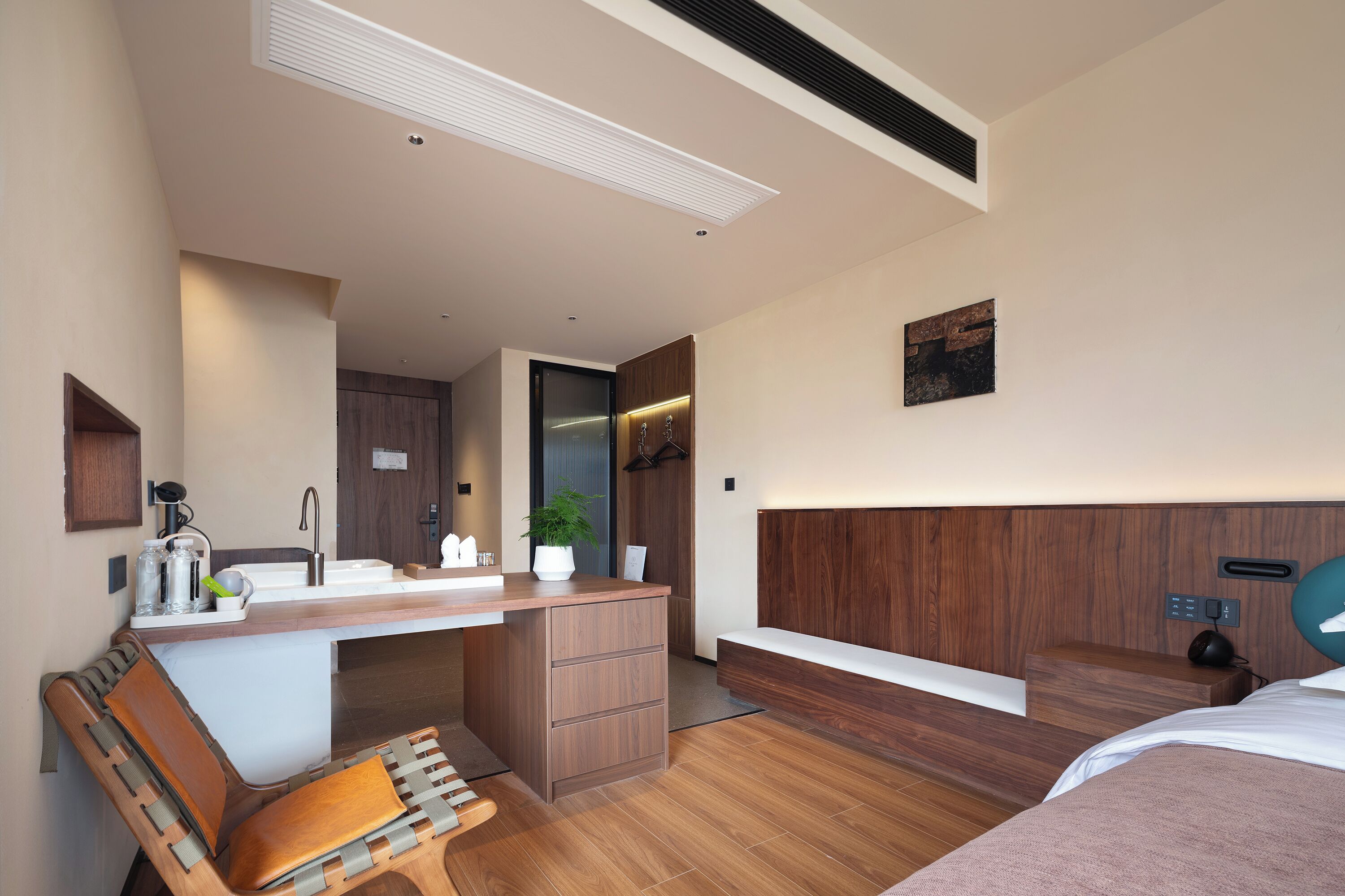 A modern hotel room with a wooden desk, a bench, a bed, and a small kitchenette.