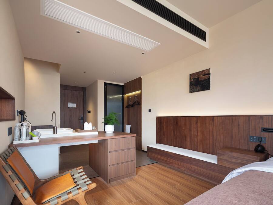 A modern hotel room with a wooden desk, a bench, a bed, and a small kitchenette.