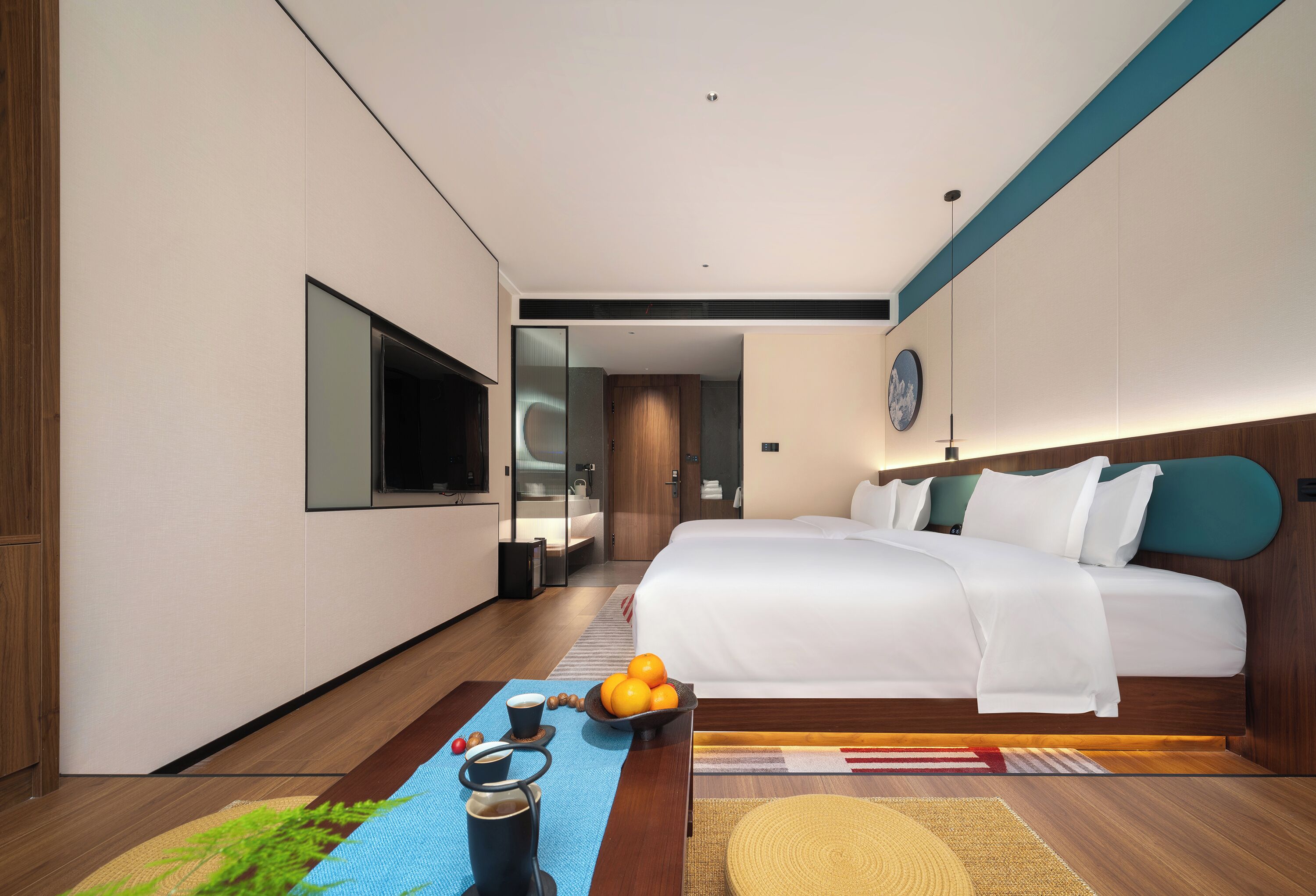 A modern hotel room with a large bed, a flat-screen TV, a coffee table with fruits and a cup, and a view into the hallway.