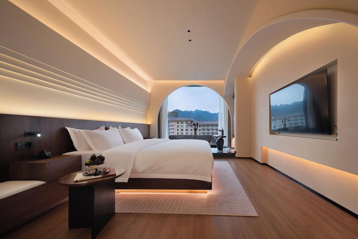 A modern hotel room with a large bed, a bedside table, a flat-screen TV mounted on the wall, and a view of a cityscape through an arched window.