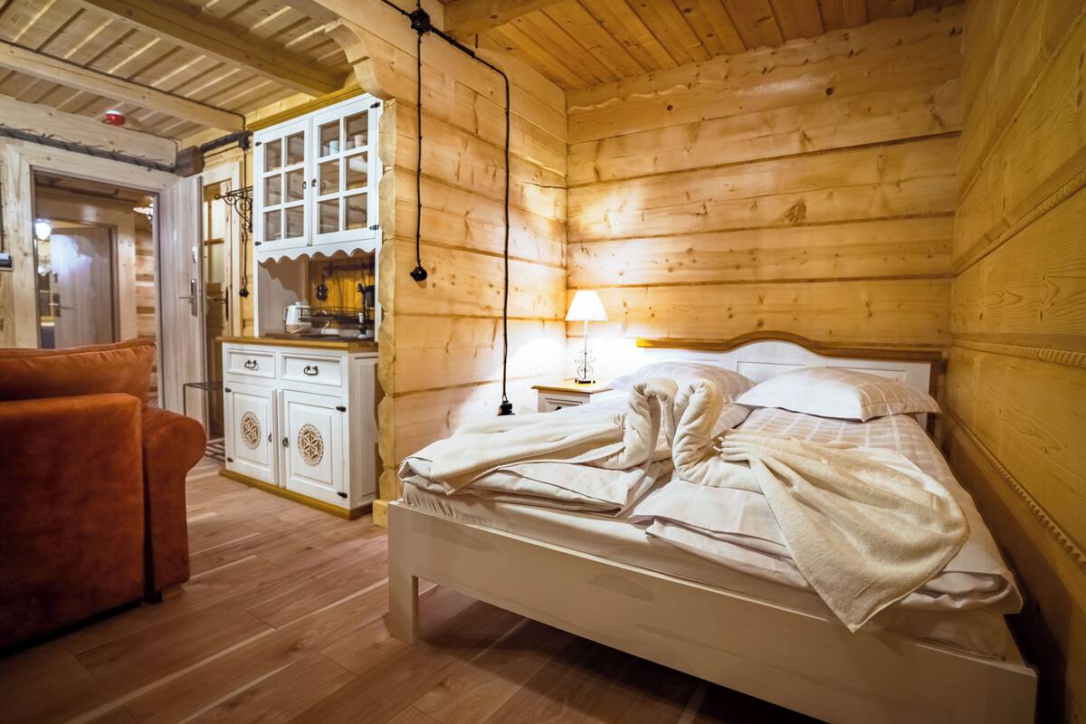 A wooden cabin bedroom with a bed, a wooden dresser, a lamp, and a sofa.