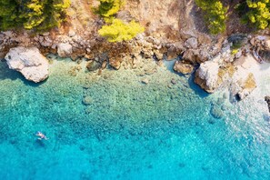 An aerial view of a coastal area with clear turquoise waters, rocky shoreline, and sparse vegetation.