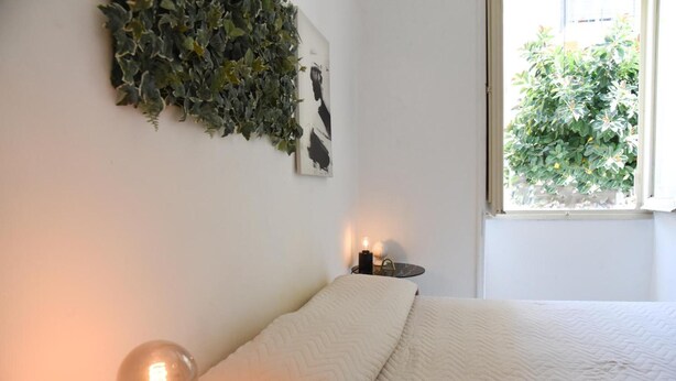 A bedroom with a bed, a wall-mounted light, a small table with a candle, and a window with a view of greenery outside. - Apartment 'Viabalduini2' with Wi-Fi and Air Conditioning (Lecce)