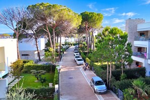 A residential street lined with trees and parked cars, leading to a building with balconies.