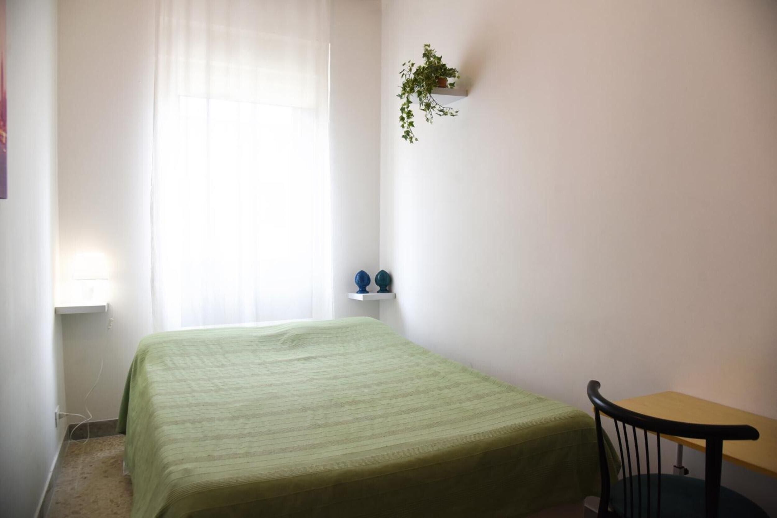 A room with a bed, a small table, a chair, a window with sheer curtains, and a wall-mounted plant.