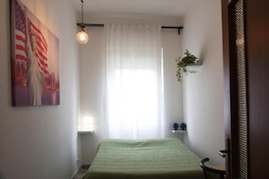A bedroom with a bed, a window with sheer curtains, a wall-mounted light fixture, and a small shelf with a plant.