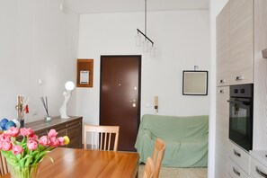A modern kitchen with a built-in oven, a dining area with a wooden table and chairs, a green-covered sofa, and a framed picture on the wall.