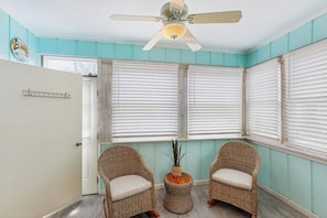 A room with teal walls, white shutters, wicker chairs, a ceiling fan, and a small table with a vase.