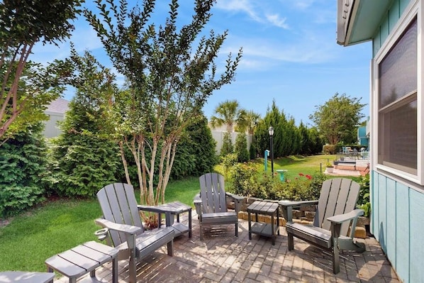 A patio with Adirondack chairs and a table, surrounded by greenery and a lawn.