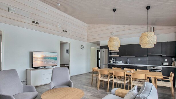 Una sala moderna con un sofá gris, una mesa de centro redonda de madera y un sillón gris. Hay una zona de cocina con armarios oscuros y un refrigerador de acero inoxidable.