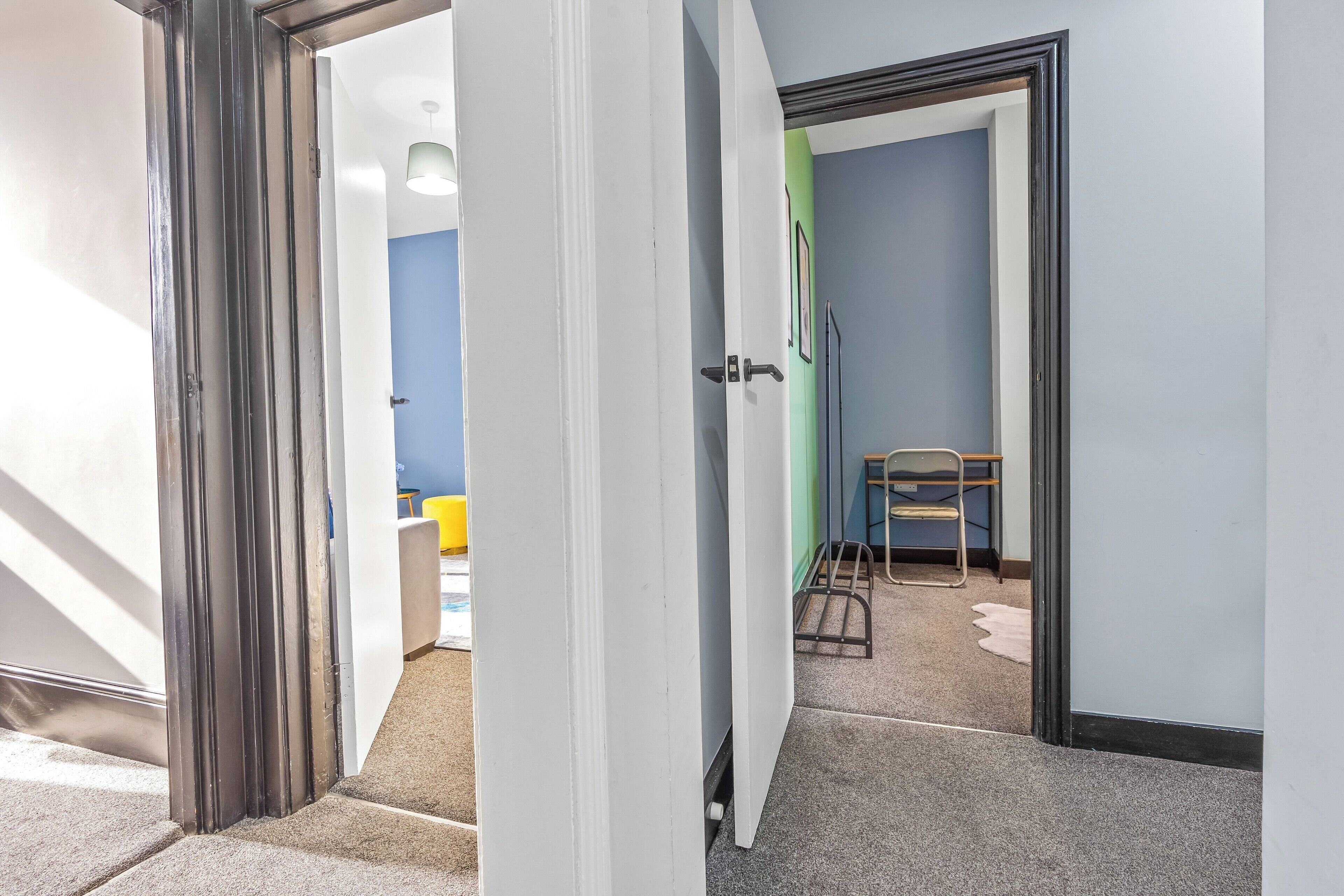 Un couloir avec une porte blanche qui donne sur une pièce dont un mur est bleu et l’autre, vert. Il y a une chaise et une petite table dans la pièce.