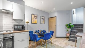 Una cocina moderna con armarios blancos, un microondas integrado y una estufa a gas. Hay una zona de comedor con una mesa redonda y sillas azules. Se observa una escalera al fondo.