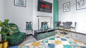 Ett modernt vardagsrum med en grön soffa, en öppen spis, ett soffbord i glas och en platt-TV som visar "NETFLIX".