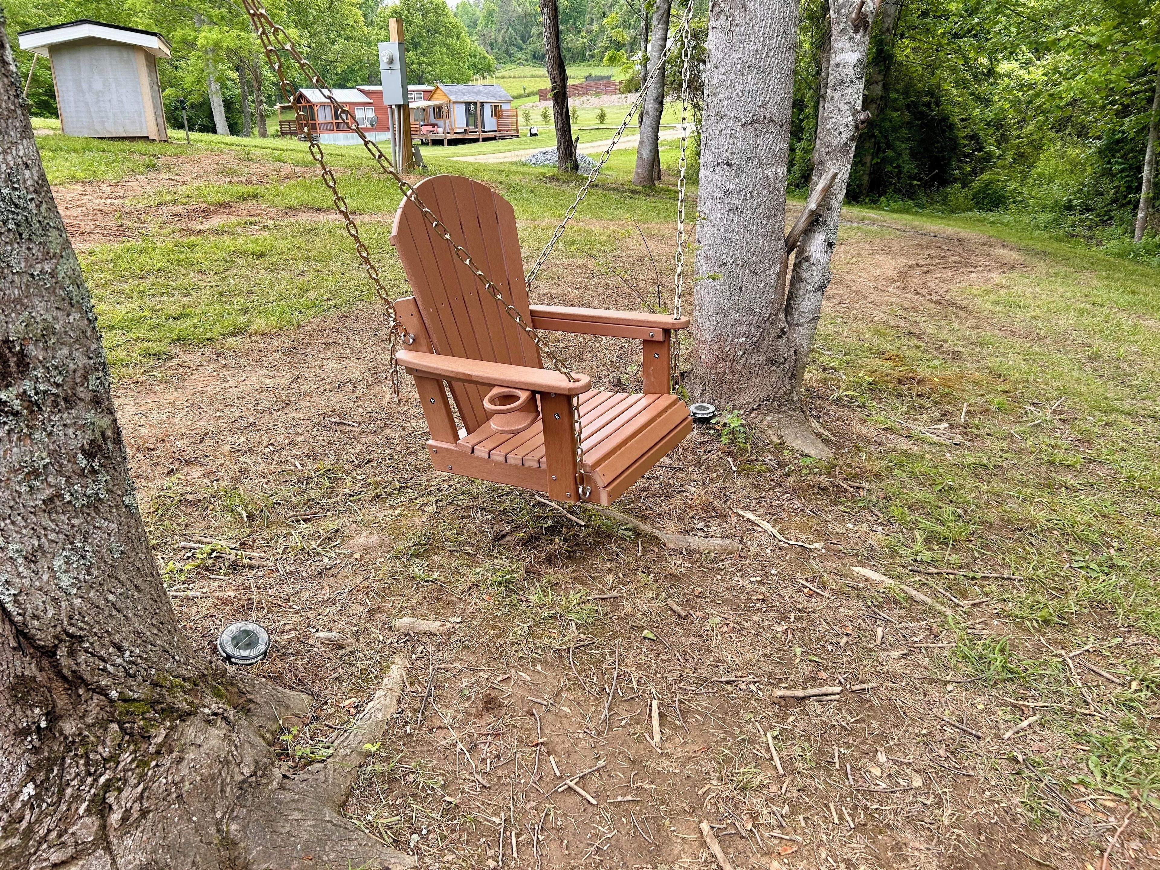 A wooden swing chair in a wooded area with a small shed and a cabin in the background.