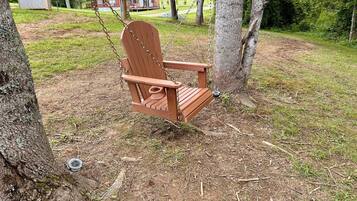 A wooden swing chair in a wooded area with a small shed and a cabin in the background.