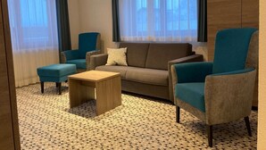 Et hotelværelse med sofa, to lænestole, et sofabord og store vinduer med gardiner.