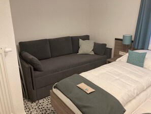 Ein Zimmer mit grauem Sofa, einem Bett mit weißer Bettwäsche, einem kleinen Tisch, einer Lampe und einem Fenster mit Vorhängen.