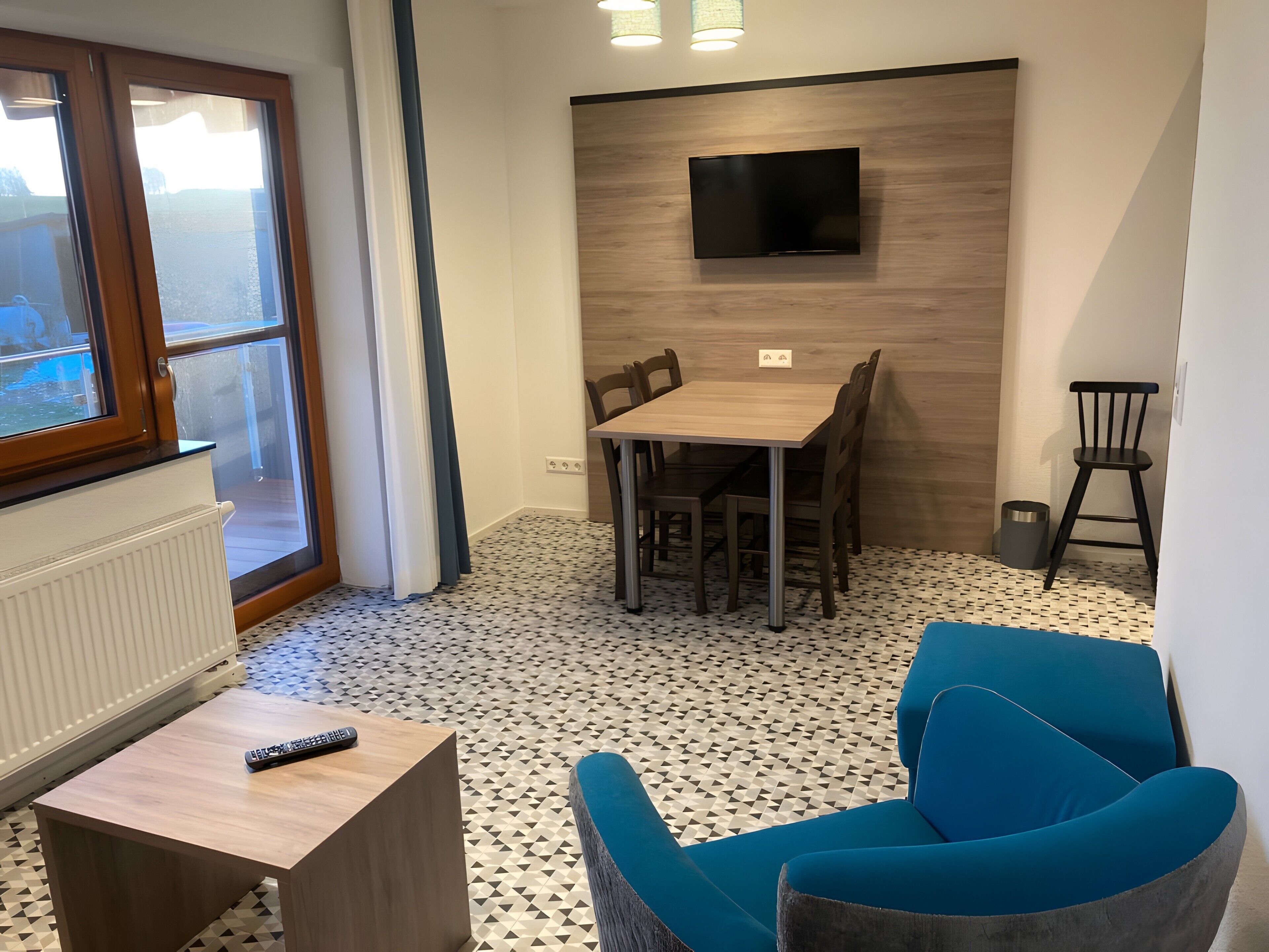 Une pièce moderne comprenant une table à manger, des chaises, un téléviseur fixé au mur, un fauteuil bleu et une petite table avec une télécommande.