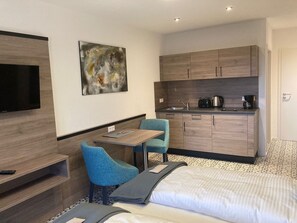 Ein Hotelzimmer mit Bett, Fernseher, einem kleinen Tisch mit Stühlen, einer Küchenzeile und einem Bild an der Wand.