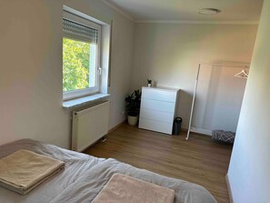 A bedroom with a bed, a dresser, a radiator, a window with blinds, a potted plant, and a hanging clothes rack.