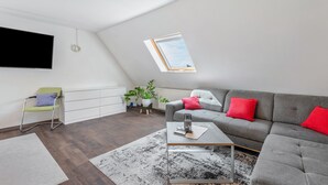 A modern living room with a sloped ceiling, a flat-screen TV, a grey sofa with red cushions, a coffee table, and a small plant.