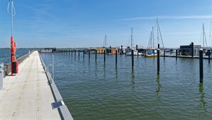 Um cais se estende sobre águas calmas, com barcos atracados, céu claro e uma linha de costa ao longe.