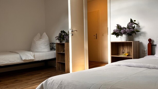 Slaapkamer met twee bedden, een houten vloer, een deur naar een andere kamer en een klein plankje met een bloevas.